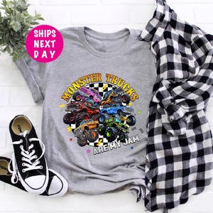 May include: A gray t-shirt featuring a colorful graphic of monster trucks with the text "MONSTER TRUCKS ARE MY JAM." The design includes various monster trucks in different colors. Black and white sneakers and a plaid shirt are also in the image.