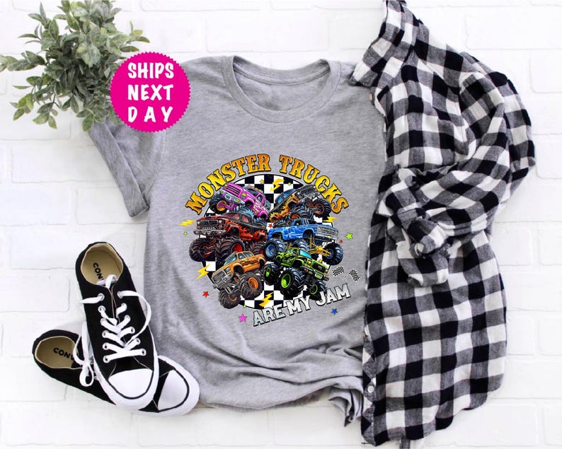 May include: A gray t-shirt featuring a colorful graphic of monster trucks with the text "MONSTER TRUCKS ARE MY JAM." The design includes various monster trucks in different colors. Black and white sneakers and a plaid shirt are also in the image.