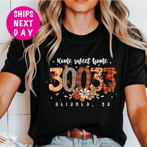 Puede incluir: Camiseta negra con la frase "Home sweet home" y los números "30033" en una fuente decorativa. El texto "Atlanta, GA" está debajo. Un círculo rosa con "SHIPS NEXT DAY" está en la esquina superior izquierda.