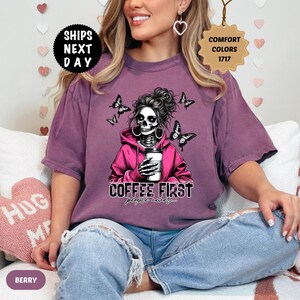 Op de afbeelding: Bessenkleurig T-shirt met een afbeelding van een schedel met een roze hoodie en een koffiekopje, met vlinders. De tekst "COFFEE FIRST people never..." staat eronder. Het shirt heeft een relaxte pasvorm.
