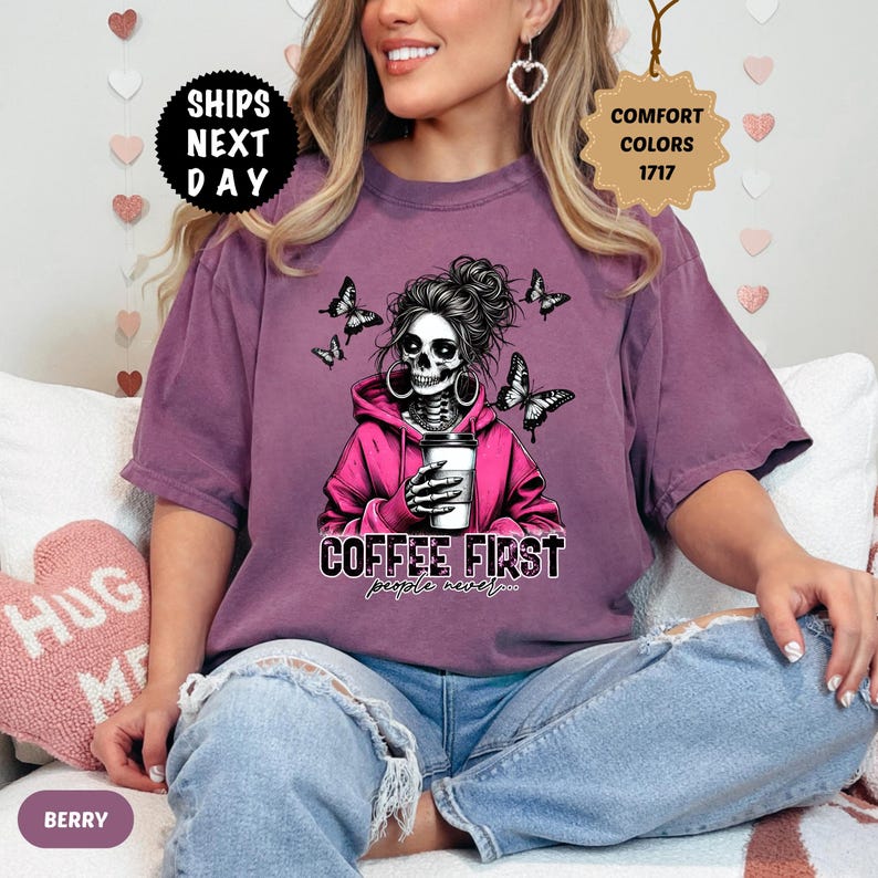 Op de afbeelding: Bessenkleurig T-shirt met een afbeelding van een schedel met een roze hoodie en een koffiekopje, met vlinders. De tekst "COFFEE FIRST people never..." staat eronder. Het shirt heeft een relaxte pasvorm.