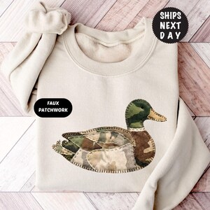 Peut inclure: Sweat-shirt beige avec un motif de canard en patchwork. Le canard a une tête verte, un corps marron et des ailes camouflage. Le sweat-shirt a un col rond et des manches longues. Le texte "FAUX PATCHWORK" est sur le côté gauche.