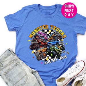 May include: A blue t-shirt featuring a colorful graphic of monster trucks arranged in a circle. The text "MONSTER TRUCKS ARE MY JAM" is displayed in a circular design. The shirt is paired with denim shorts and white sneakers.