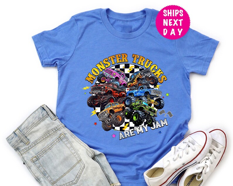 May include: A blue t-shirt featuring a colorful graphic of monster trucks arranged in a circle. The text "MONSTER TRUCKS ARE MY JAM" is displayed in a circular design. The shirt is paired with denim shorts and white sneakers.