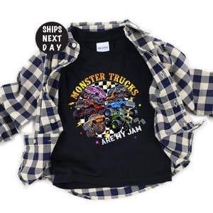 May include: Black t-shirt with a colorful graphic of monster trucks and the text "MONSTER TRUCKS ARE MY JAM". The shirt is layered with a blue and white plaid button-down shirt.