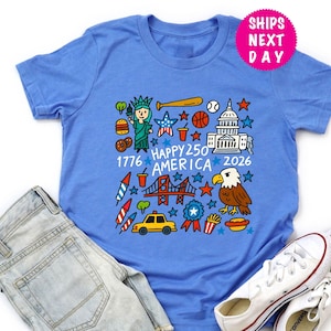 Puede incluir: Una camiseta azul claro con un gr&aacute;fico colorido que presenta iconos estadounidenses como la Estatua de la Libertad, un bate de b&eacute;isbol y el Capitolio. Tambi&eacute;n incluye el texto "Happy 250 America 2026".