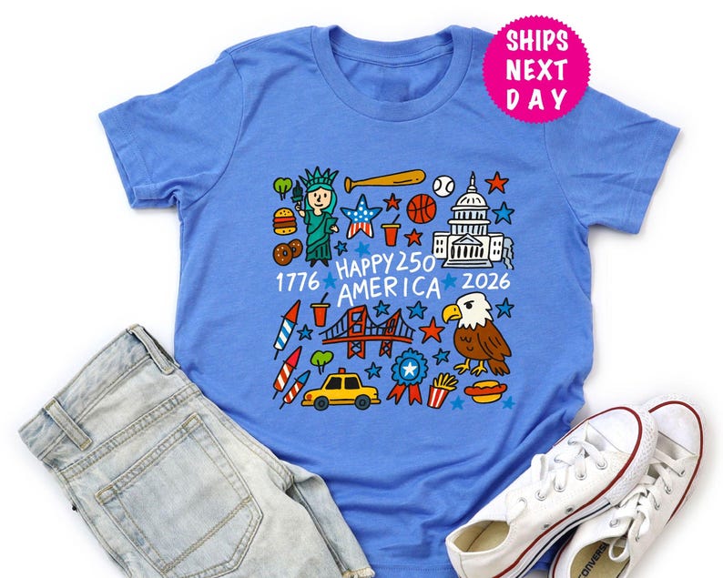 Puede incluir: Una camiseta azul claro con un gr&aacute;fico colorido que presenta iconos estadounidenses como la Estatua de la Libertad, un bate de b&eacute;isbol y el Capitolio. Tambi&eacute;n incluye el texto "Happy 250 America 2026".