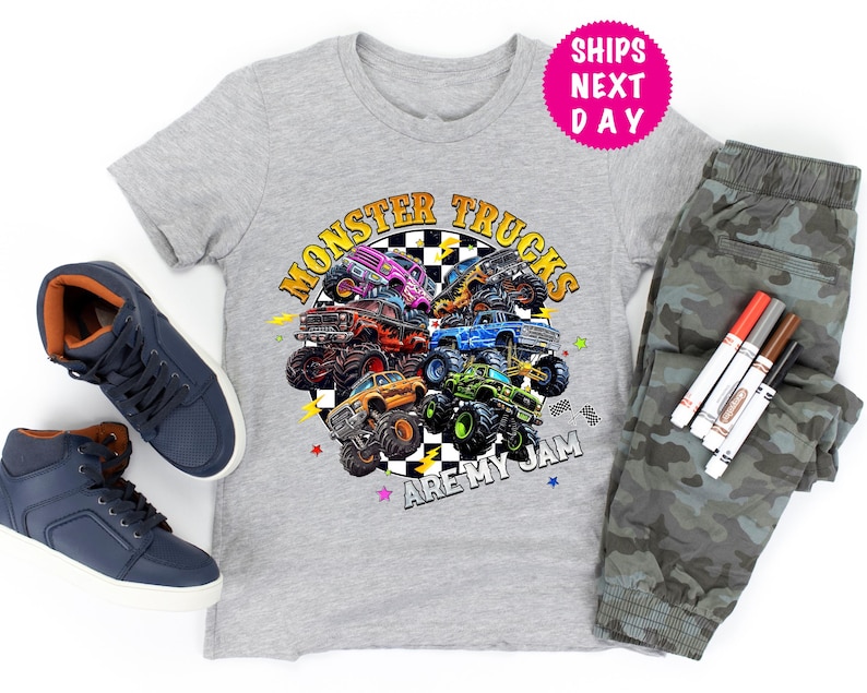 May include: A gray t-shirt with a colorful graphic of monster trucks and the text "Monster Trucks Are My Jam." The image also includes a pair of blue sneakers, camouflage pants, and markers.