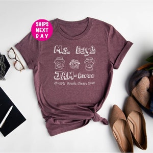 May include: A maroon t-shirt with white text that reads "Ms. Day's JAM-berec Sixth Grade Class, 2023". The text is stylized to look like a handwritten note. There are three jars of jam drawn above the text.