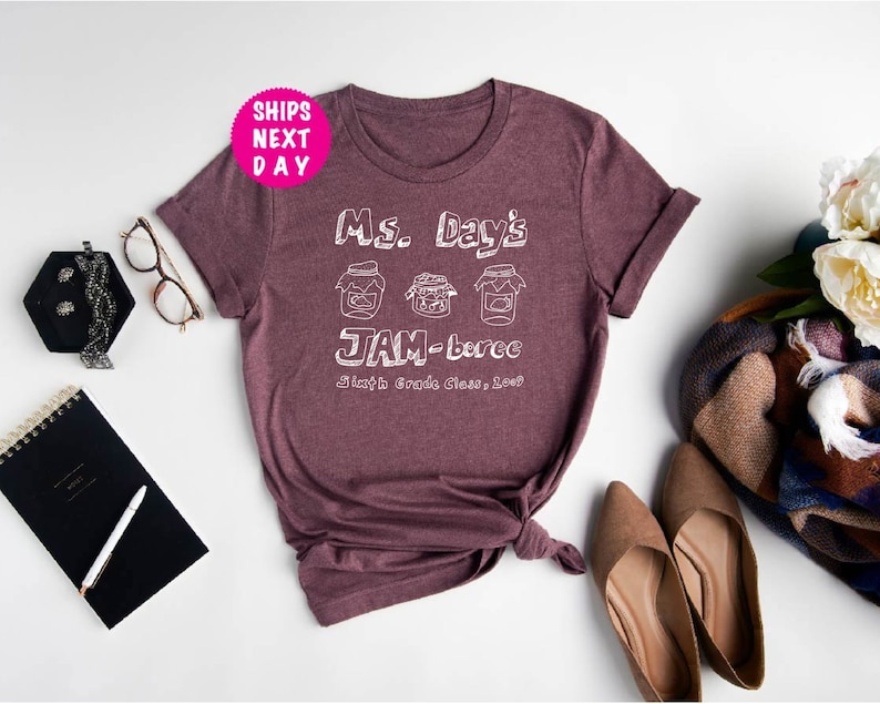May include: A maroon t-shirt with white text that reads "Ms. Day's JAM-berec Sixth Grade Class, 2023". The text is stylized to look like a handwritten note. There are three jars of jam drawn above the text.
