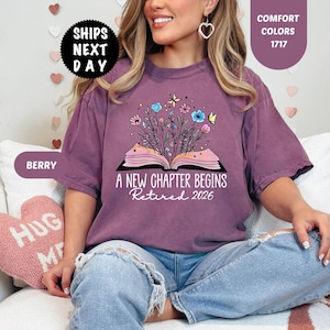 May include: Berry-colored t-shirt with the text "A NEW CHAPTER BEGINS Retired 2026" and a graphic of an open book with flowers and butterflies. The shirt has short sleeves and a crew neck.