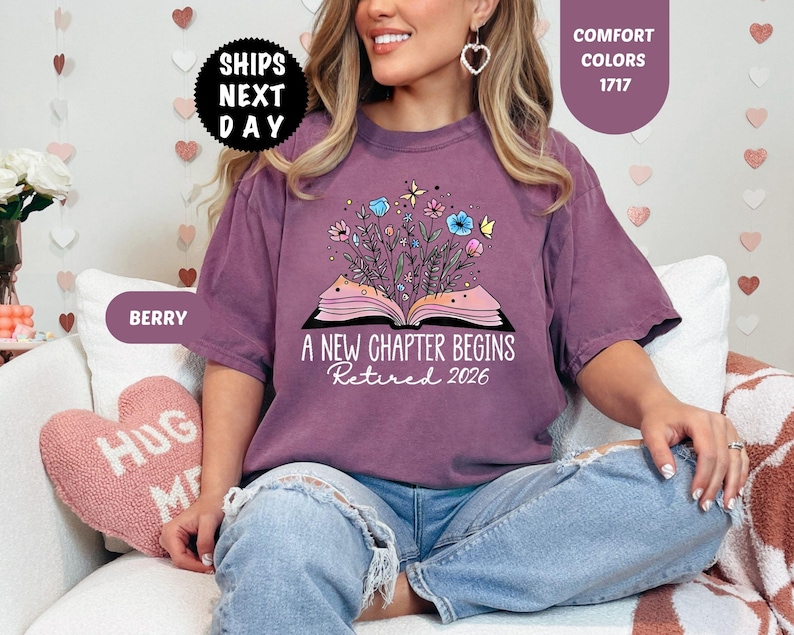 May include: Berry-colored t-shirt with the text "A NEW CHAPTER BEGINS Retired 2026" and a graphic of an open book with flowers and butterflies. The shirt has short sleeves and a crew neck.