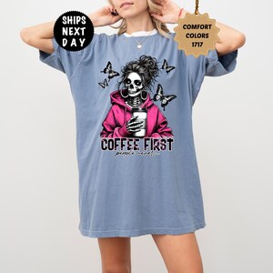 Op de afbeelding: Lichtblauw T-shirt met een afbeelding van een schedel met een roze hoodie en een koffiekopje. De tekst "COFFEE FIRST people never" staat eronder. Vlinders zijn ook afgebeeld in het ontwerp.