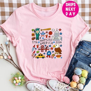 Puede incluir: Una camiseta rosa claro con un gr&aacute;fico colorido que celebra el 250 aniversario de Am&eacute;rica. El dise&ntilde;o incluye la Estatua de la Libertad, un bate de b&eacute;isbol, el Capitolio, un &aacute;guila y el texto "Happy 250 America 2026".
