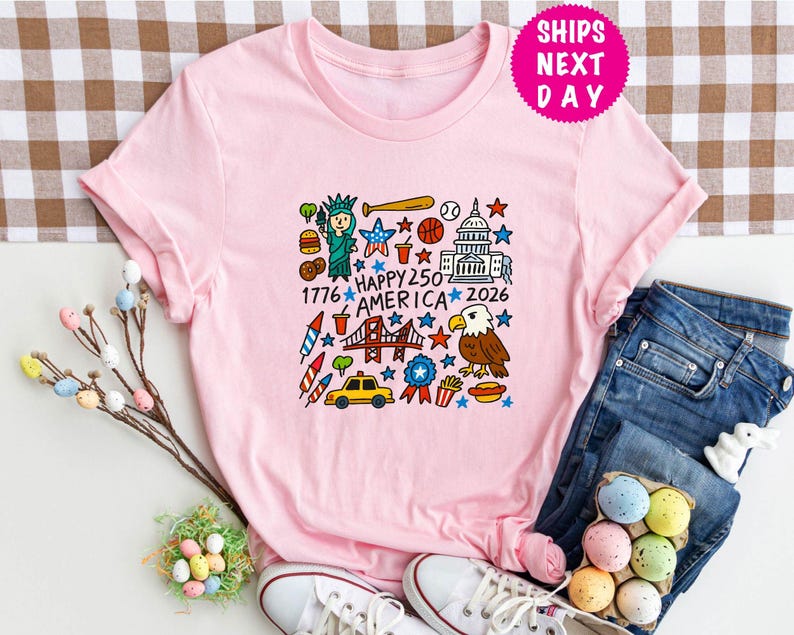 Puede incluir: Una camiseta rosa claro con un gr&aacute;fico colorido que celebra el 250 aniversario de Am&eacute;rica. El dise&ntilde;o incluye la Estatua de la Libertad, un bate de b&eacute;isbol, el Capitolio, un &aacute;guila y el texto "Happy 250 America 2026".