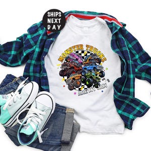 May include: White t-shirt with a colorful graphic of monster trucks and the text "Monster Trucks Are My Jam." The shirt is paired with a blue and green plaid shirt, blue jeans, and mint green and white sneakers.