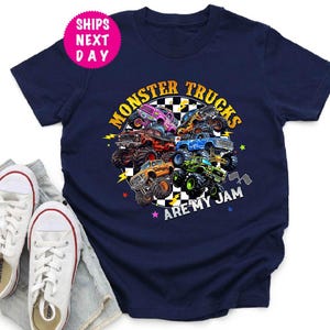 May include: Navy blue t-shirt featuring a colorful graphic of monster trucks with the text "Monster Trucks Are My Jam." The design includes various monster trucks in different colors. White canvas sneakers and denim shorts are in the foreground.