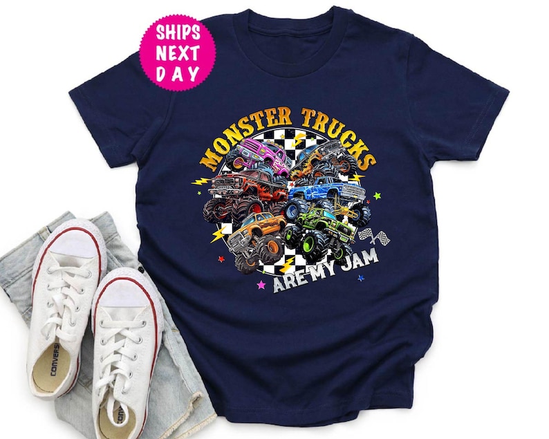May include: Navy blue t-shirt featuring a colorful graphic of monster trucks with the text "Monster Trucks Are My Jam." The design includes various monster trucks in different colors. White canvas sneakers and denim shorts are in the foreground.