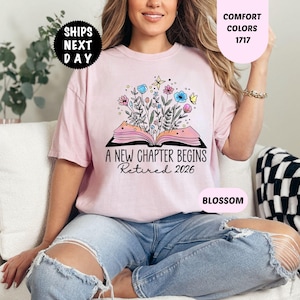 May include: A light pink t-shirt with the text "A New Chapter Begins Retired 2026" and a floral design. The shirt is a Comfort Colors 1717 in Blossom. The image also includes a "Ships Next Day" graphic.