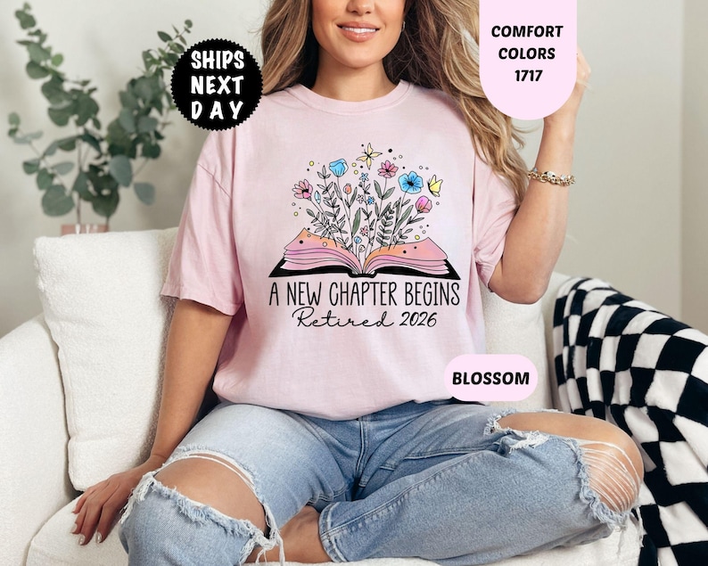 May include: A light pink t-shirt with the text "A New Chapter Begins Retired 2026" and a floral design. The shirt is a Comfort Colors 1717 in Blossom. The image also includes a "Ships Next Day" graphic.