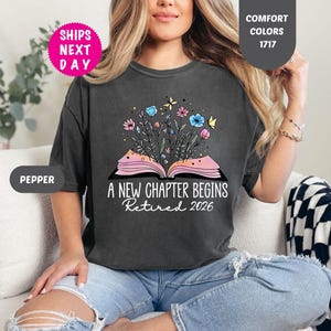 May include: Dark gray t-shirt with the text "A New Chapter Begins Retired 2026". The shirt features a graphic of an open book with flowers and butterflies. The shirt is a Comfort Colors 1717.