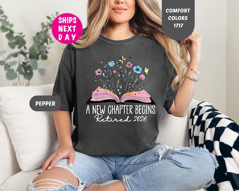 May include: Dark gray t-shirt with the text "A New Chapter Begins Retired 2026". The shirt features a graphic of an open book with flowers and butterflies. The shirt is a Comfort Colors 1717.