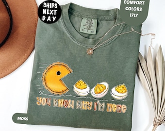 May include: A moss green crew neck t-shirt featuring a graphic of a pumpkin pie slice chasing three deviled eggs. The text below the graphic reads, "YOU KNOW WHY I'M HERE." The shirt has a Comfort Colors brand label on the inside collar.