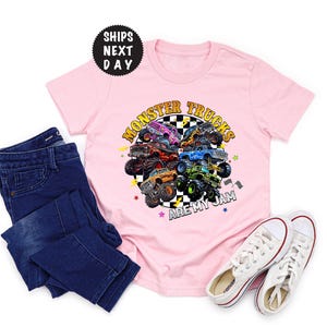 May include: A light pink t-shirt with a colorful graphic of monster trucks in a circle, with the text "Monster Trucks Are My Jam." The shirt is paired with dark blue jeans and white sneakers.