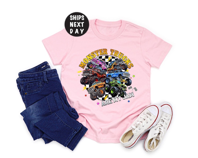 May include: A light pink t-shirt with a colorful graphic of monster trucks in a circle, with the text "Monster Trucks Are My Jam." The shirt is paired with dark blue jeans and white sneakers.
