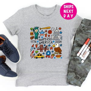 Puede incluir: Una camiseta gris con un gr&aacute;fico colorido que celebra el 250 aniversario de Am&eacute;rica, con la Estatua de la Libertad, un bate de b&eacute;isbol y el Capitolio. La imagen incluye zapatillas azules y pantalones de camuflaje.