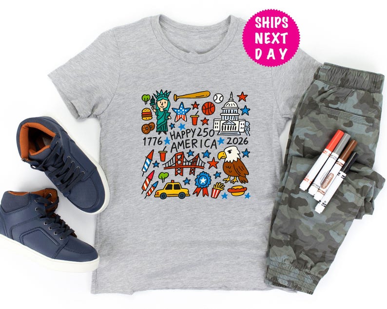 Puede incluir: Una camiseta gris con un gr&aacute;fico colorido que celebra el 250 aniversario de Am&eacute;rica, con la Estatua de la Libertad, un bate de b&eacute;isbol y el Capitolio. La imagen incluye zapatillas azules y pantalones de camuflaje.