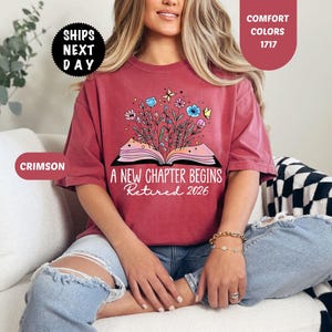 May include: Crimson-colored t-shirt with the text "A New Chapter Begins Retired 2026" and a graphic of an open book with flowers and butterflies. The shirt is a comfort color.