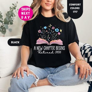 May include: Black t-shirt with the text "A NEW CHAPTER BEGINS Retired 2026" and a graphic of an open book with flowers. The shirt has a round neckline and short sleeves. The shirt is part of the "Comfort Colors" collection.