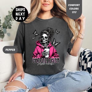 Op de afbeelding: Donkergrijs t-shirt met een afbeelding van een schedel met een roze hoodie en een koffiekopje. De tekst "COFFEE FIRST people never..." staat eronder. Zwarte vlinders omringen de schedel.