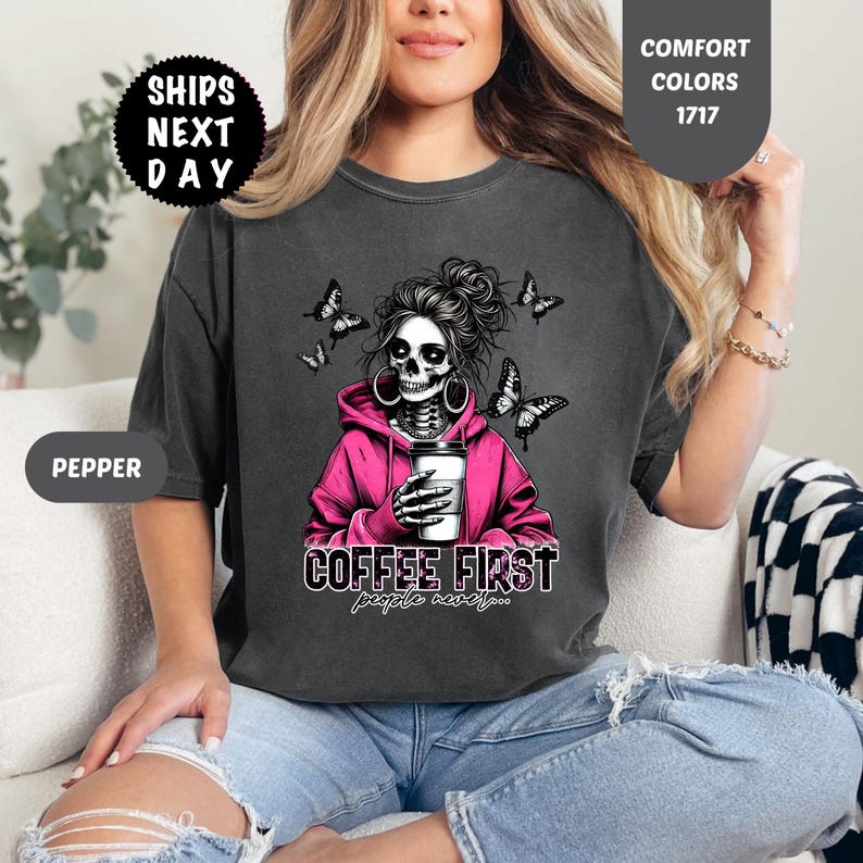 Op de afbeelding: Donkergrijs t-shirt met een afbeelding van een schedel met een roze hoodie en een koffiekopje. De tekst "COFFEE FIRST people never..." staat eronder. Zwarte vlinders omringen de schedel.