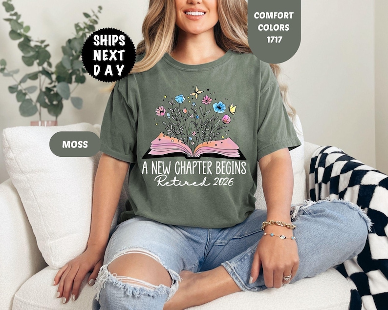 May include: Moss green t-shirt with the text "A NEW CHAPTER BEGINS Retired 2026". The graphic features an open book with flowers and butterflies. The shirt is a comfort color.