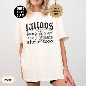 May include: An ivory short-sleeve t-shirt featuring black text that reads: tattoos because life is hard AND I DESERVE stickers. The design includes a checkered pattern at the end of the word stickers and a circular graphic around the word life.