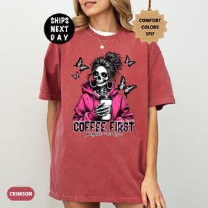 Op de afbeelding: Karmizijnrood T-shirt met een afbeelding van een schedel met een roze hoodie die een koffiekopje vasthoudt, omringd door vlinders. De tekst "COFFEE FIRST people never..." staat eronder. Onderdeel van de Comfort Colors 1717 collectie.