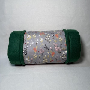 May include: A green fleece-lined cylindrical pillow with a floral print fabric. The fabric features a light gray background with a variety of small flowers in shades of pink, yellow, and orange.