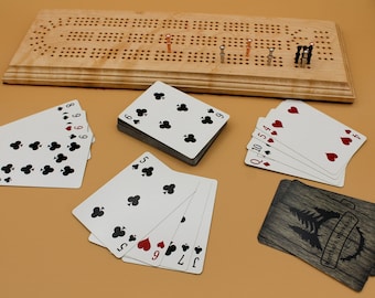 May include: A wooden cribbage board with metal pegs in copper and silver tones, accompanied by several playing cards. The cards include a deck, a hand of clubs, a hand of hearts, and a hand of mixed suits. One card back features a tree graphic.