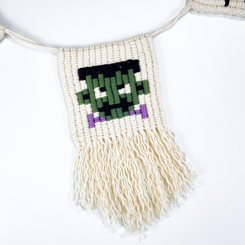 May include: A white string banner with a green, black, and purple pixelated Frankenstein face. The banner has white fringe on the bottom.