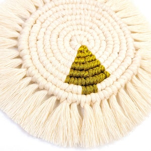 May include: A round, white macrame coaster with a gold triangle in the center. The coaster has a fringe border.
