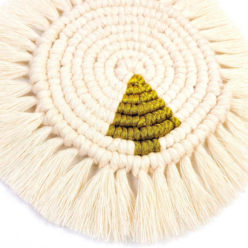 May include: A round, white macrame coaster with a gold triangle in the center. The coaster has a fringe border.