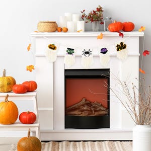 May include: White fireplace mantel with a Halloween-themed knitted banner featuring a ghost, a monster, a spider, a witch's hat, and a bat. The banner is hanging above a black electric fireplace with a realistic log display. There are pumpkins and fall decorations on the mantel.