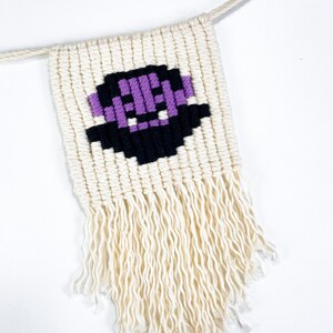 May include: A white macrame wall hanging with a purple and black pixelated design. The hanging has a fringe at the bottom.