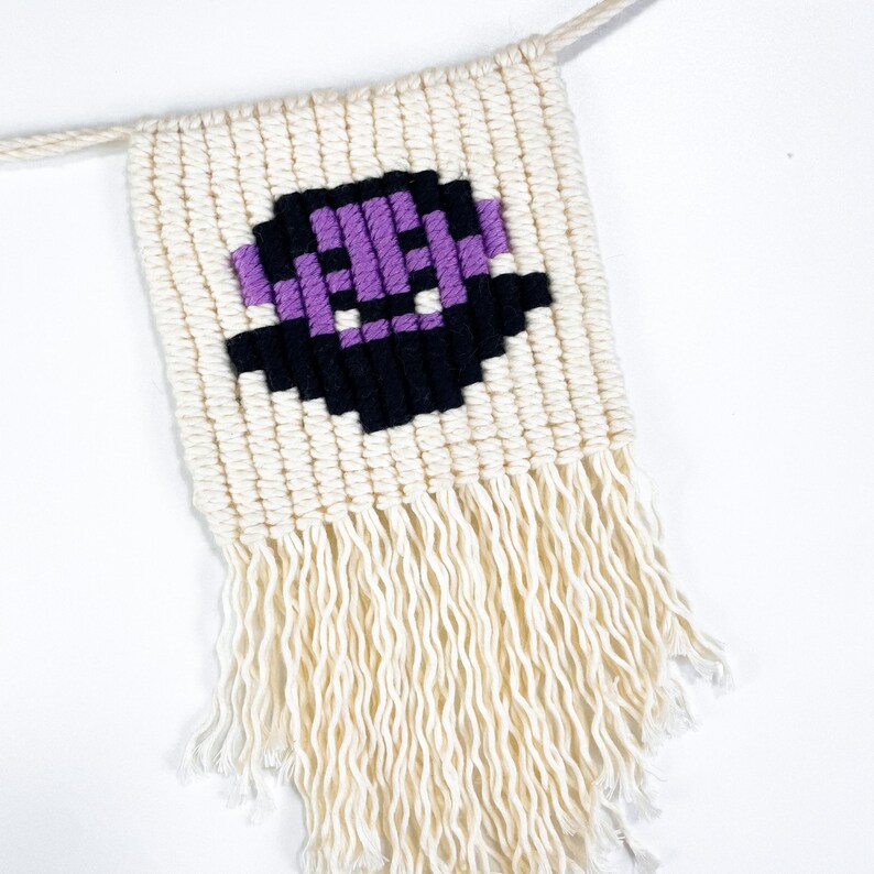 May include: A white macrame wall hanging with a purple and black pixelated design. The hanging has a fringe at the bottom.