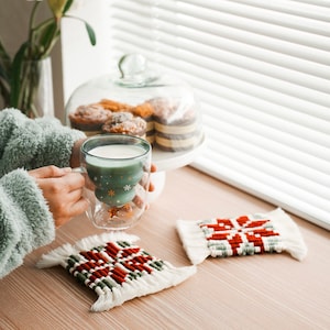 May include: A green glass mug with a snowflake design holds a warm beverage. Two woven coasters with a red and white snowflake design are on a wooden table.