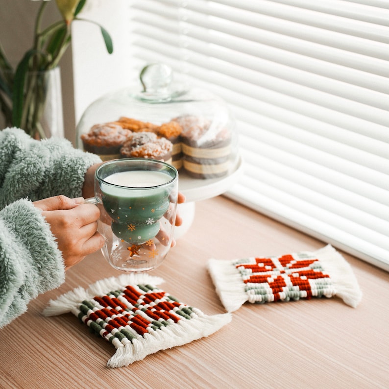May include: A green glass mug with a snowflake design holds a warm beverage. Two woven coasters with a red and white snowflake design are on a wooden table.