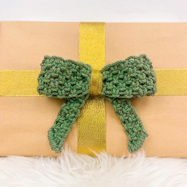 K&ouml;nnte beinhalten: Eine braune Geschenkbox mit einer gr&uuml;nen geh&auml;kelten Schleife, die oben gebunden ist. Die Box ist mit goldener Schleife umwickelt.