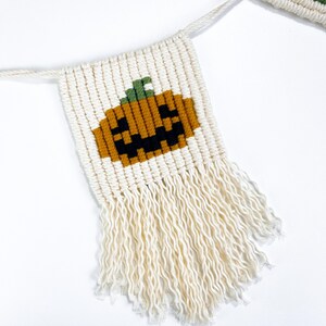 May include: A white macrame banner with a brown and black pumpkin face design. The banner has white fringe on the bottom.